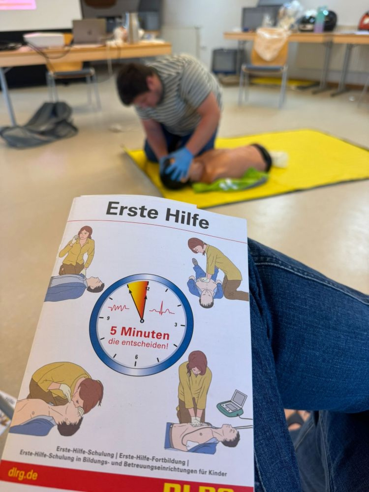 Im Vordergrund ein Flyer eines Erste-Hilfe Kurses und im Hintergrund eine Person beim Üben von Reanimationsmaßnahmen
